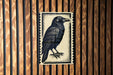 a black and white illustration of a bird, specifically a crow, mounted on a wooden wall.