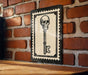 A framed image of a skull with a key is displayed on a brick wall.