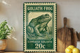 A green and white postage stamp featuring a detailed illustration of a frog, with the text "GOLIATH FROG" and "CONRUA GOLIATH" printed on it. The stamp is placed on a wooden surface, with a potted plant, a wooden cutting board, and some lemons nearby.