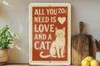 a sign with a cat and the text "All you need is love and a cat" on a shelf.