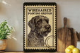 a framed illustration of a wirehaired pointing griffin on a shelf, with a wooden cutting board and a potted plant nearby.