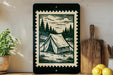a framed illustration of a tent set up in a forest, surrounded by pine trees and a cloudy sky, placed on a shelf next to a wooden cutting board and a potted plant.