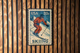 A vintage 20c US postage stamp featuring a skier in a red suit and helmet, mounted on a wooden wall.