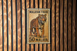 A metal stamp with a tiger image and the text "MALAYAN TIGER 50 MALAYA" is displayed on a wooden wall.