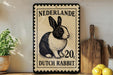 A decorative sign featuring a black and white illustration of a Dutch rabbit, with the text "Nederlande 20, Dutch Rabbit" printed on it. The sign is placed on a wooden surface, with a potted plant, a wooden cutting board, and some lemons nearby.