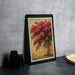 A framed painting of a vibrant floral arrangement is displayed on a dark surface, with a stack of black camera lenses and a white dish visible to the side.
