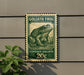 A green and white postage stamp featuring a detailed illustration of a frog, mounted on a wall next to a plant.