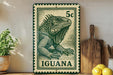 A green and white postage stamp featuring an illustration of an iguana, with the word "Iguana" printed on it. The stamp is placed on a shelf next to a wooden cutting board and some potted plants.