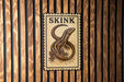 A sign with the word "SKINK" and an illustration of a lizard or salamander is displayed on a wooden wall.