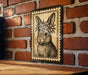 A framed illustration of a rabbit wearing a flower crown is displayed on a brick wall.