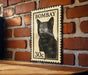 A vintage-style metal stamp featuring a black cat on a brick wall.