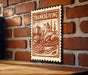 A framed Thanksgiving-themed stamp is displayed on a brick wall, featuring an illustration of a table set for a meal with various dishes and utensils.