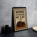 A metal sign with the text "HOME IS WHERE MY CAT IS" and an illustration of a cat is placed on a countertop next to some black camera lenses.