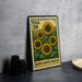 A metal stamp with a vibrant illustration of sunflowers on a green background, placed on a gray countertop next to some black camera lenses.