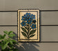 A decorative tile with a blue flower design is mounted on a wall, accompanied by a small green plant to the left.