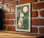 A framed illustration of a rabbit holding a balloon is displayed on a brick wall.