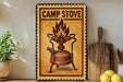 a sign that reads "CAMP STOVE" with an illustration of a camp stove on it. The sign is placed on a shelf next to a wooden cutting board and a potted plant.