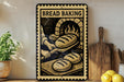 A vintage-style metal sign with the text "BREAD BAKING" and an illustration of a baker working in a brick oven. The sign is displayed on a wooden shelf, with a potted plant, wooden cutting board, and lemons nearby.