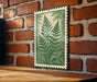A green and white framed print of a fern plant is displayed on a brick wall, with a wooden table below it.