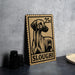 A wooden postcard featuring a dog illustration, with the text "SLOUGHI" printed on it, is displayed on a table next to some black camera lenses.