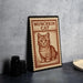 A framed illustration of a cat is displayed on a table, with a stack of black containers and a woven basket visible in the background.
