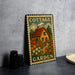 A metal sign with a cottage garden design, featuring a house surrounded by a garden with flowers, is displayed on a table.