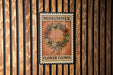 A vintage-style metal sign with the text "MIDSUMMER FLOWER CROWN" displayed on it, mounted on a wooden wall.