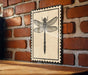 A framed image of a dragonfly is displayed on a brick wall.