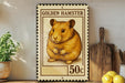 A framed stamp depicting a golden hamster, with the text "GOLDEN HAMSTER" and "50c" printed on it. The stamp is placed on a wooden surface, with a potted plant, a wooden cutting board, and some lemons nearby.