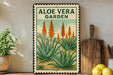 a framed illustration of an aloe vera plant, with the text "ALOE VERA GARDEN" prominently displayed. The frame is placed on a wooden surface, and there are some potted plants and kitchen utensils visible in the background.