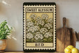 a framed print of a flower with the text "Sweet Alyssum BED" on it, placed on a shelf next to a wooden cutting board and some potted plants.