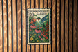A colorful postage stamp featuring a hummingbird surrounded by flowers and plants, hanging on a wooden wall.