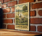 A framed illustration of a lamb standing in a grassy field, with a tree in the background, is mounted on a brick wall.