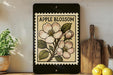 a framed illustration of an apple blossom on a shelf, with a wooden cutting board and a potted plant to the left.