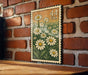 A framed print of a daisy flower design is hanging on a brick wall.