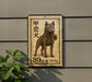 A metal sign with a stamp-like design featuring a dog, mounted on a wall next to a plant.