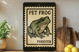 A framed image of a green frog with the text "PET FROG FOREVER" on it, placed on a shelf next to a wooden cutting board and a potted plant.