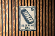 A sleeping bag postage stamp is displayed on a wooden wall.