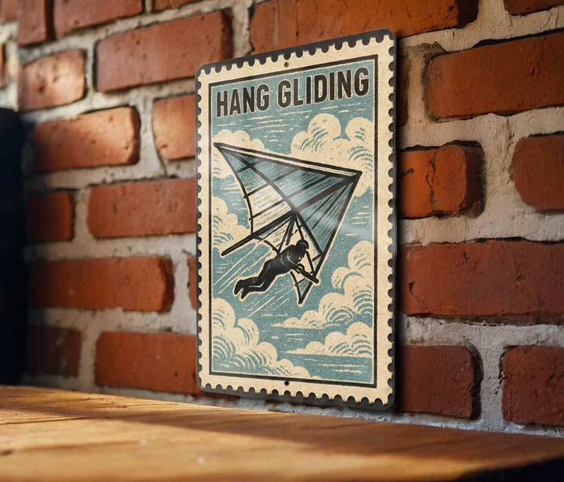 A vintage-style metal sign with the text "HANG GLIDING" and an illustration of a person hang gliding against a brick wall.