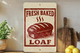 a sign that reads "Fresh Baked Loaf" with a loaf of bread depicted on it. The sign is placed on a shelf next to a wooden cutting board and a potted plant.