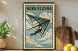 A vintage-style metal sign with the text "HANG GLIDING" and an illustration of a person hang gliding, placed on a wooden surface next to a potted plant and a wooden cutting board.