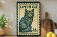 A framed vintage postage stamp featuring a cat illustration, placed on a wooden shelf next to a wooden cutting board and some lemons.
