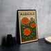 A metal stamp with a floral design and the word "MARIGOLD" printed on it, placed on a gray countertop next to some black camera lenses.