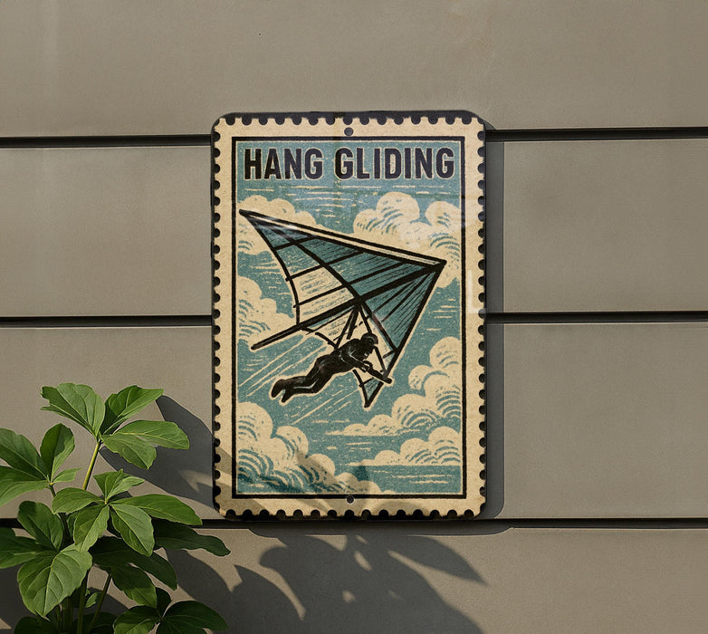 A vintage-style metal sign with the text "HANG GLIDING" and an illustration of a person hang gliding, mounted on a wall next to a plant.
