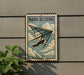 A vintage-style metal sign with the text "HANG GLIDING" and an illustration of a person hang gliding, mounted on a wall next to a plant.