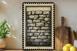 a framed artwork depicting a brick wall, a potted plant, wooden cutting boards, and a bowl of lemons on a shelf.