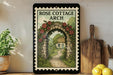 A framed illustration of a stone archway leading to a garden with a house, surrounded by greenery and flowers, is displayed on a shelf.