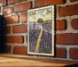 A framed illustration of a field of lavender flowers is mounted on a brick wall.