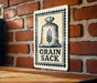 A metal sign with a graphic of a bag of grain and the text "GRAIN SACK" is displayed on a brick wall.