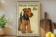 A Welsh terrier dog is depicted on a postage stamp-like sign, which is placed on a wooden surface next to a potted plant and a wooden cutting board.